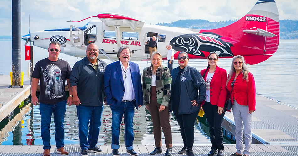 Puyallup tribal council standing on a dock with the new Kenmore Air sea plane on docked behind them. From left to right James Rideout, Fred Dillon, Bill Sterud, Sylvia Miller, Anna Bean, Annette Bryan, and Monica Miller.